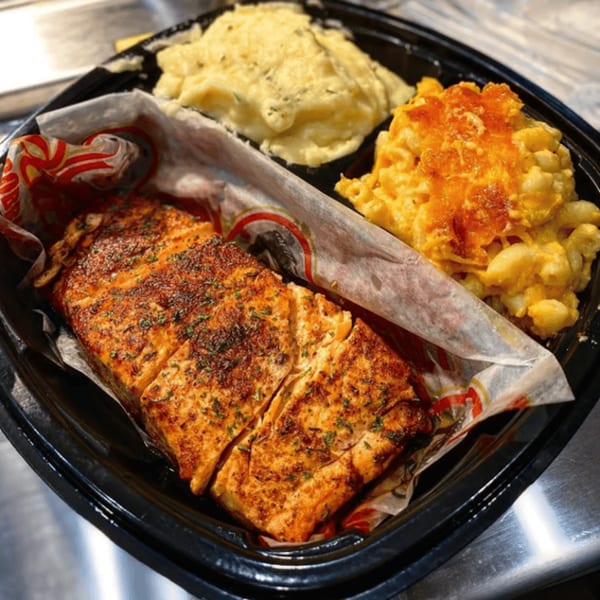 A soul food meal with baked salmon, mashed potatoes, and macaroni and cheese in a black to-go container.
