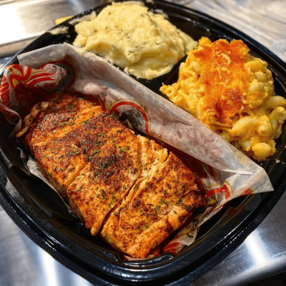 A soul food meal with baked salmon, mashed potatoes, and macaroni and cheese in a black to-go container.